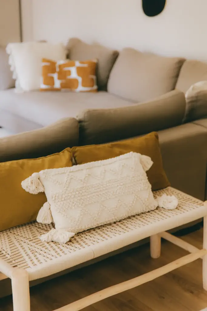 Banc en cannage au pied d’un canapé taupe, garni d’un coussin écru à pompons et de deux coussins moutarde
