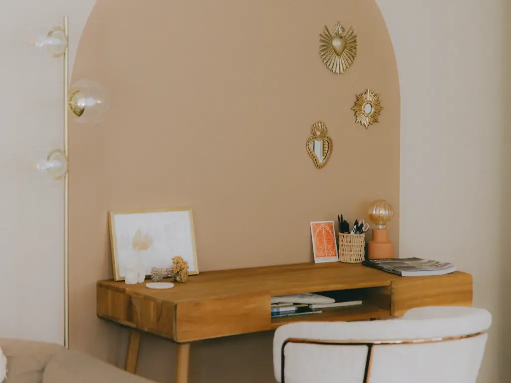 Petit bureau en bois clair sous une arche peinte couleur terre rosée, une chaise blanche bouclée, des ex-voto dorés au mur et un lampadaire à globes