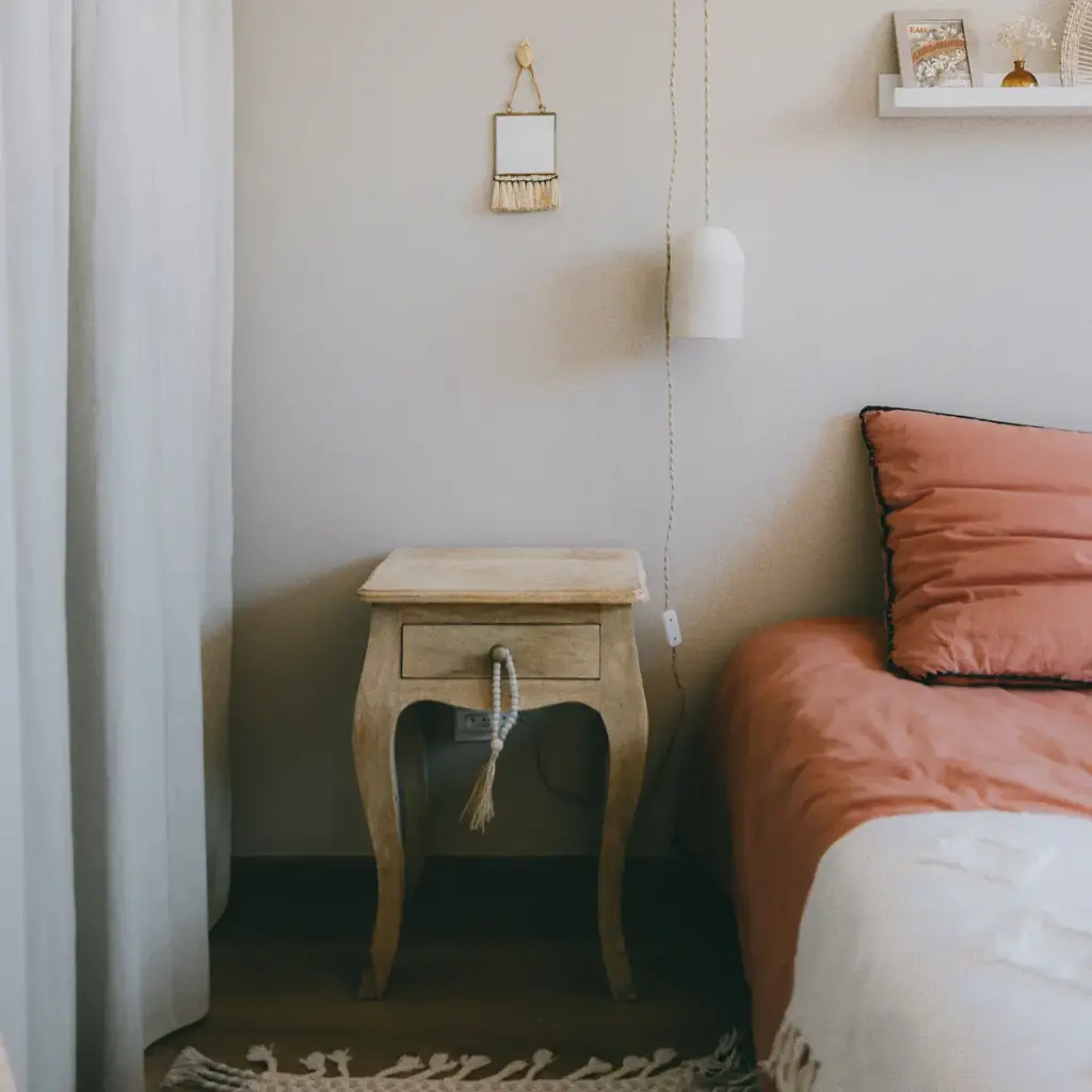 Chambre aux murs clairs, chevet en bois patiné, lampe blanche suspendue par son fil et linge de lit couleur terre cuite