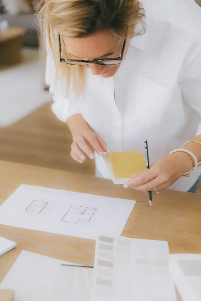 Amandine Ferreira penchée sur une table en chêne, un échantillon jaune et un crayon à la main, au-dessus d’un plan de maison et d’un nuancier