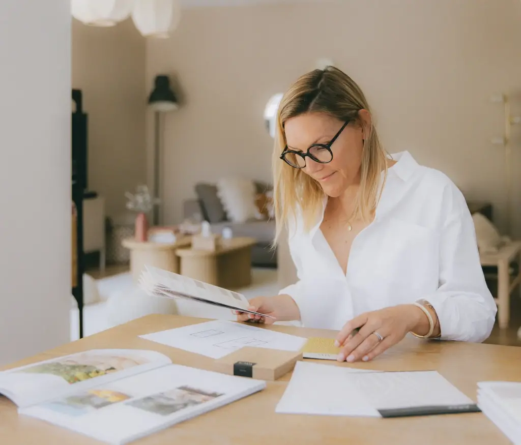 Amandine Ferreira à une grande table en bois, un nuancier à la main, penchée sur des plans, des échantillons et des livres de décoration