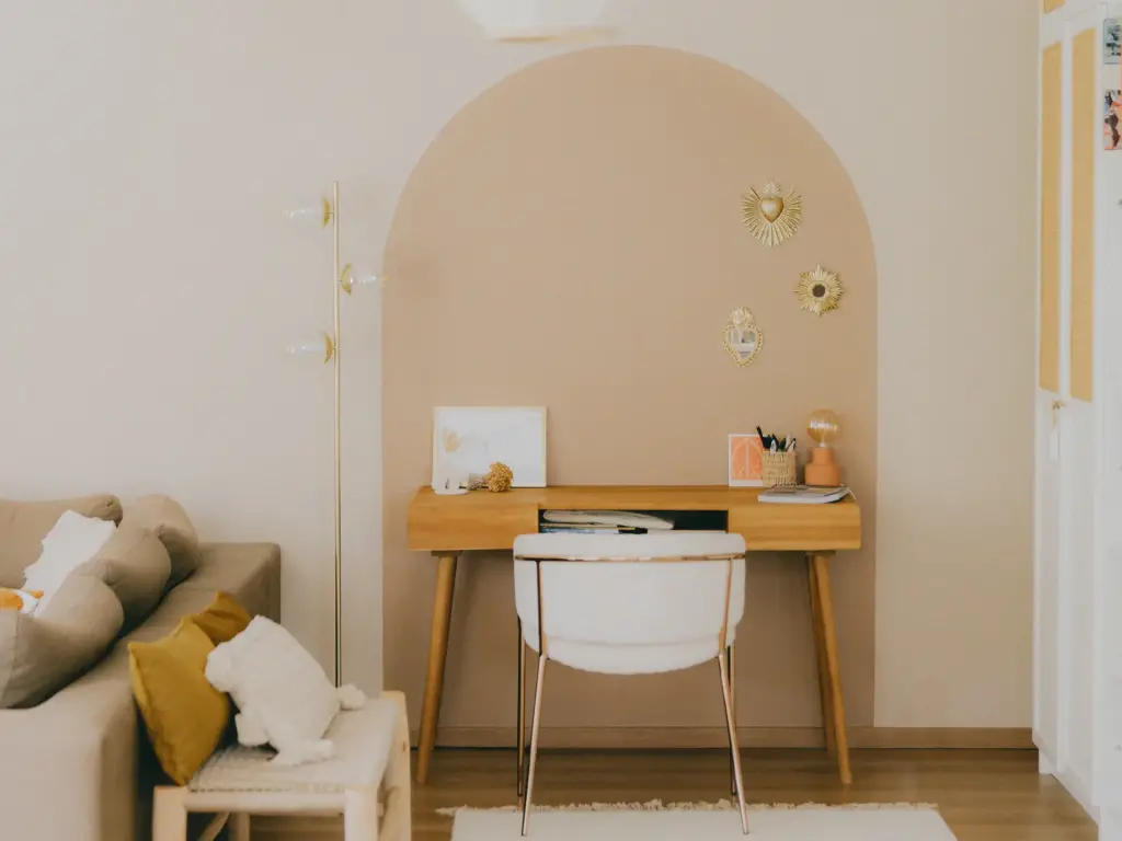 Coin bureau sous une arche peinte en rose poudré, avec un bureau en bois clair, une chaise blanche et des ex-voto dorés, près d’un canapé beige et d’un banc en cannage