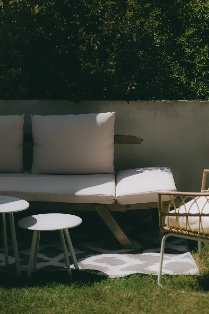 Salon de jardin : canapé en bois aux coussins écrus, petites tables rondes et fauteuil devant une haie