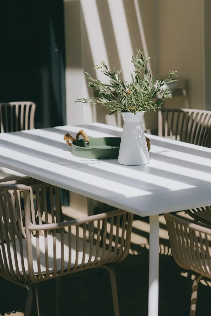 Table extérieure blanche, chaises en métal beige et vase de branches d’olivier au soleil